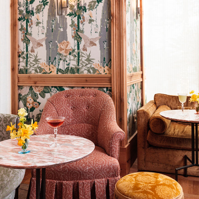 Cozy lounge corner with patterned armchairs, marble café tables, floral arrangements, and cocktails set against decorative botanical wallpaper