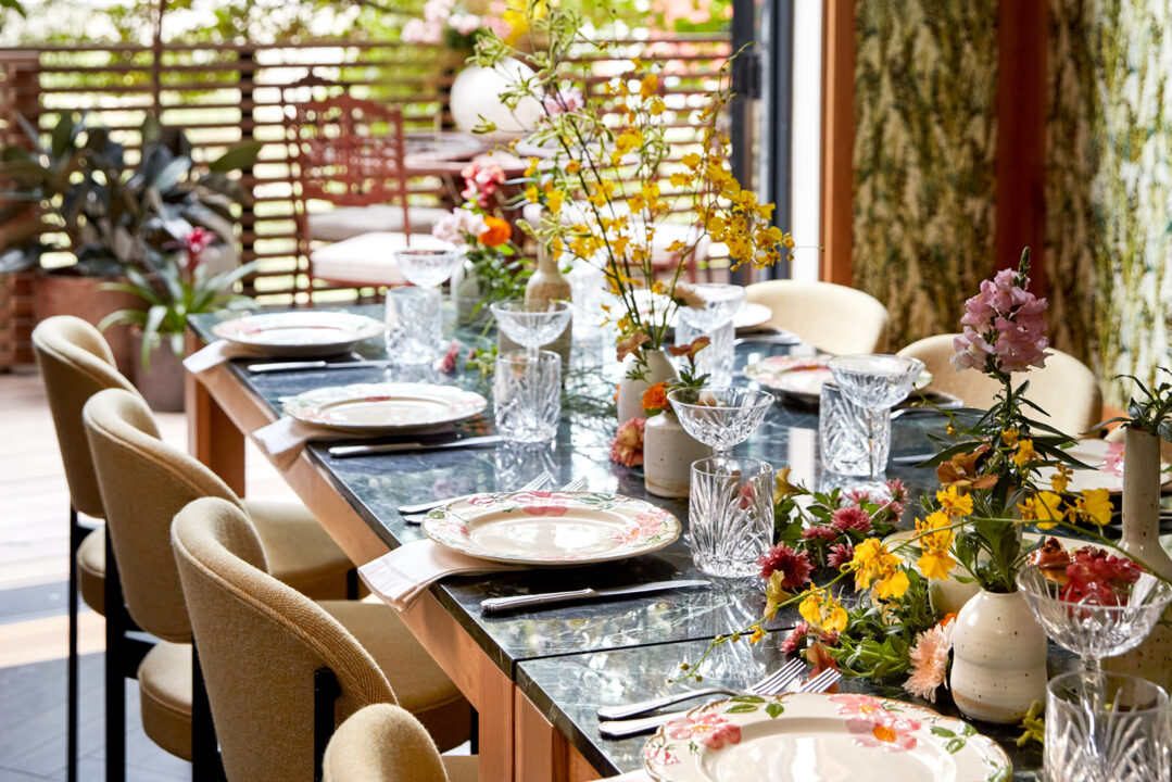 Intimate dining setup with floral arrangements, patterned plates, and elegant tableware in a sunlit space