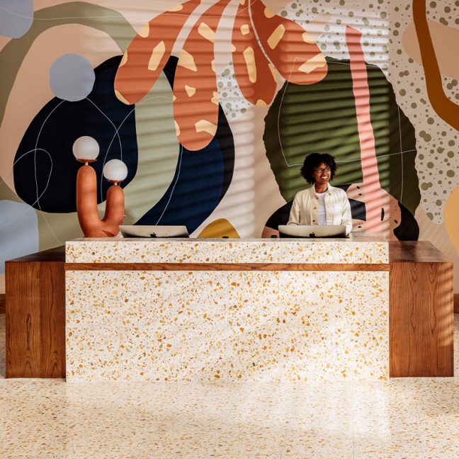 Woman behind front desk, smiling in front of Alex Proba’s Hotel June Mural