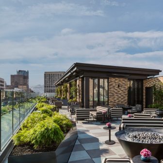 Rooftop terrace with striped lounge seating, fire pit, and city skyline views under a blue sky