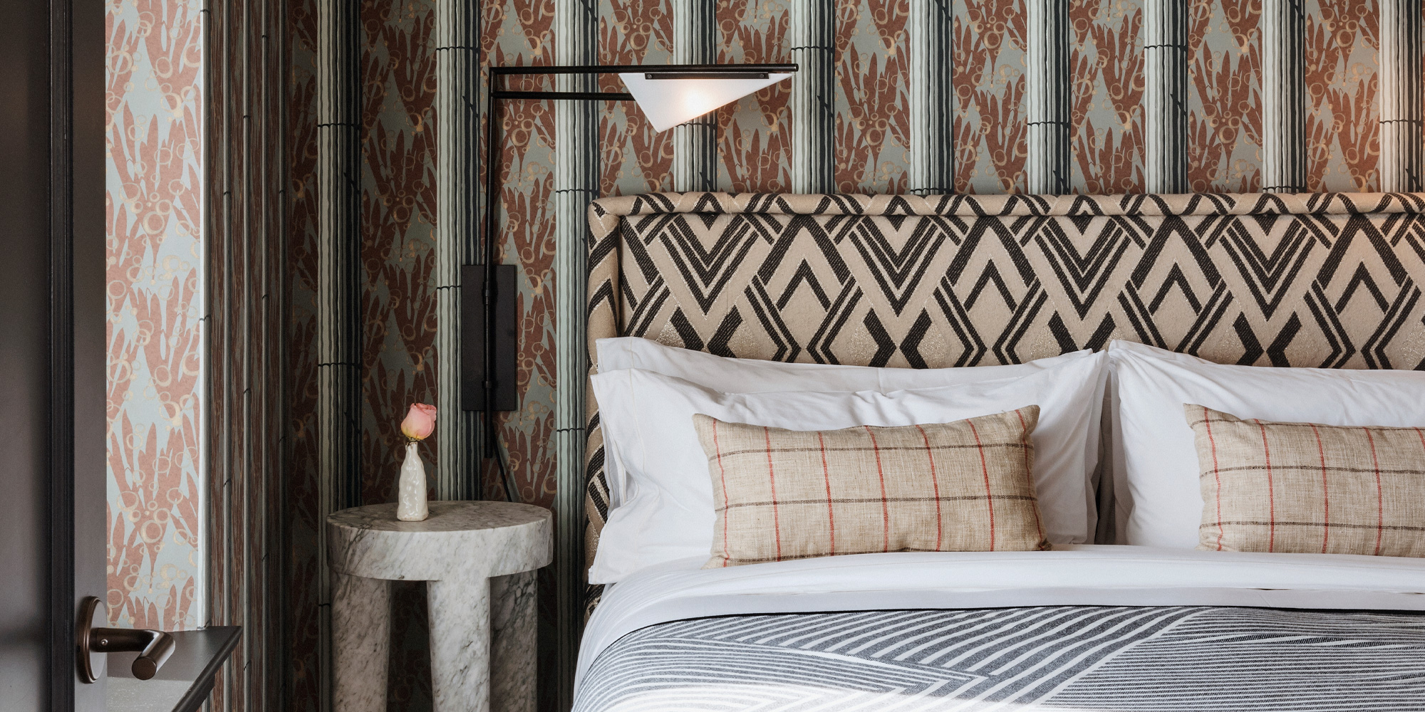 A guest room bed against clashing patterns, a black-and-cream diamond-print headboard on botanical striped wallpaper, with a swing-arm reading lamp over a small marble side table.