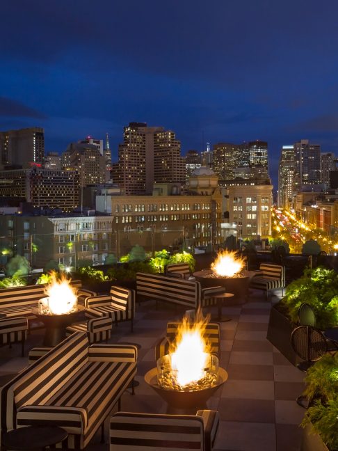 Rooftop lounge with fire pits and striped seating overlooking a city skyline at night