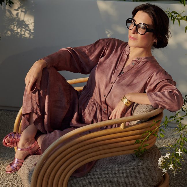 Woman wearing glasses relaxing in warm sunlight, seated against a neutral wall