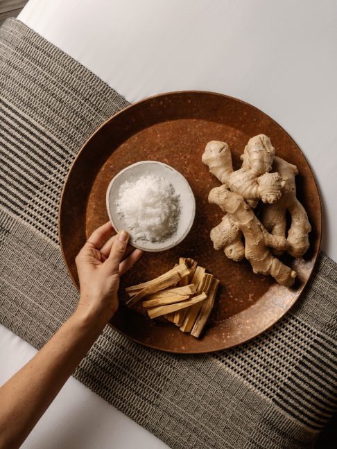Hand holding a bowl of bath salts beside ginger root and wooden sticks on a tray over a woven mat