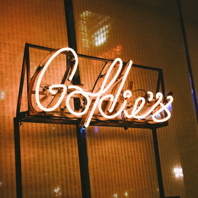 Neon “Goldie’s” sign glowing against a textured wall in a dimly lit interior