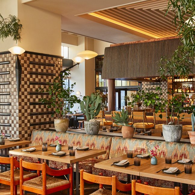 Warm, plant-filled restaurant interior with wooden tables, patterned tiles, and bar seating in the background
