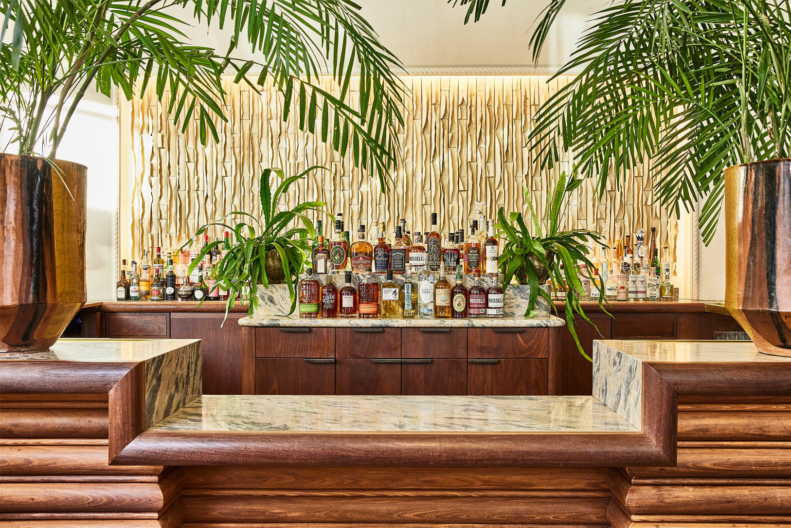 Elegant hotel bar with curated spirits display, tropical greenery, marble accents, and warm wood finishes