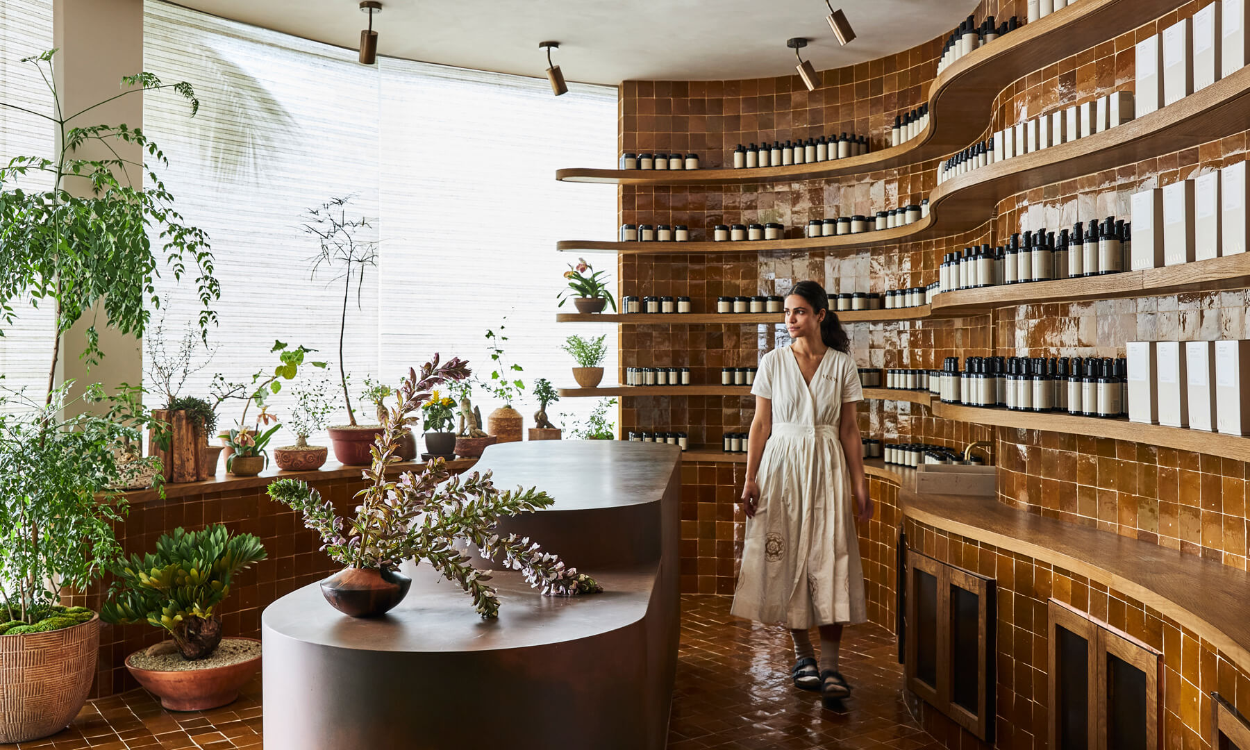 Spa retail space with curved wooden shelves of skincare products, indoor plants, and a woman walking beside a central display table