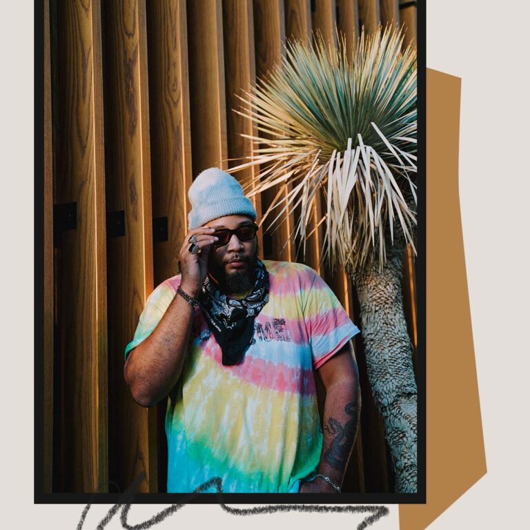 Man in tie-dye shirt and beanie standing near wooden wall and plant