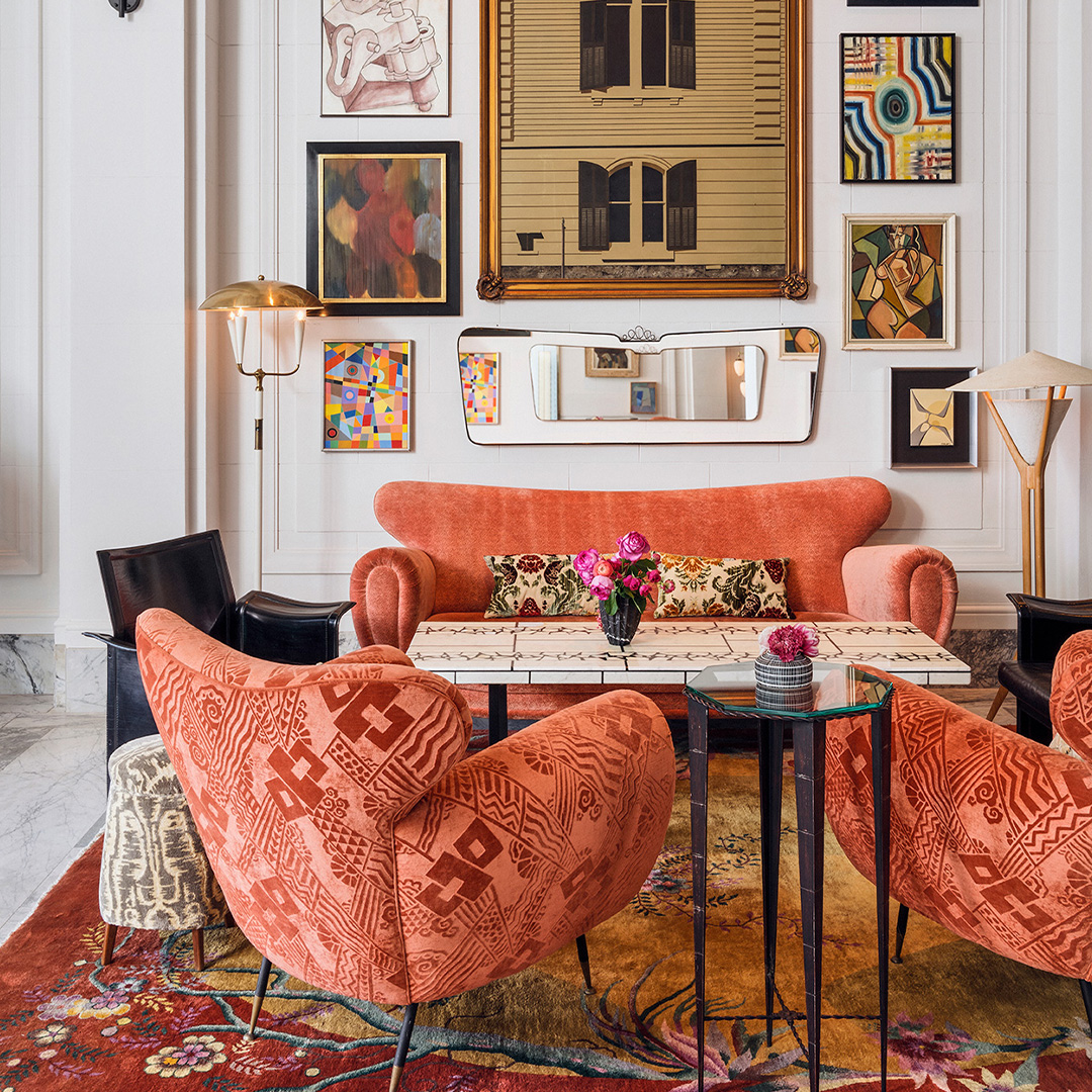 The lobby salon, coral velvet sofas and armchairs around low tables on layered vintage rugs, beneath a salon-style wall of paintings crowded frame to frame.