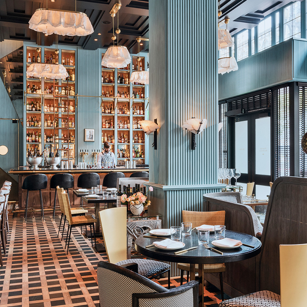 The dining room at Villon, ribbed sea-green columns and a tall backlit wall of bottles behind the bar, with round black tables set for dinner on a woven chequerboard floor.