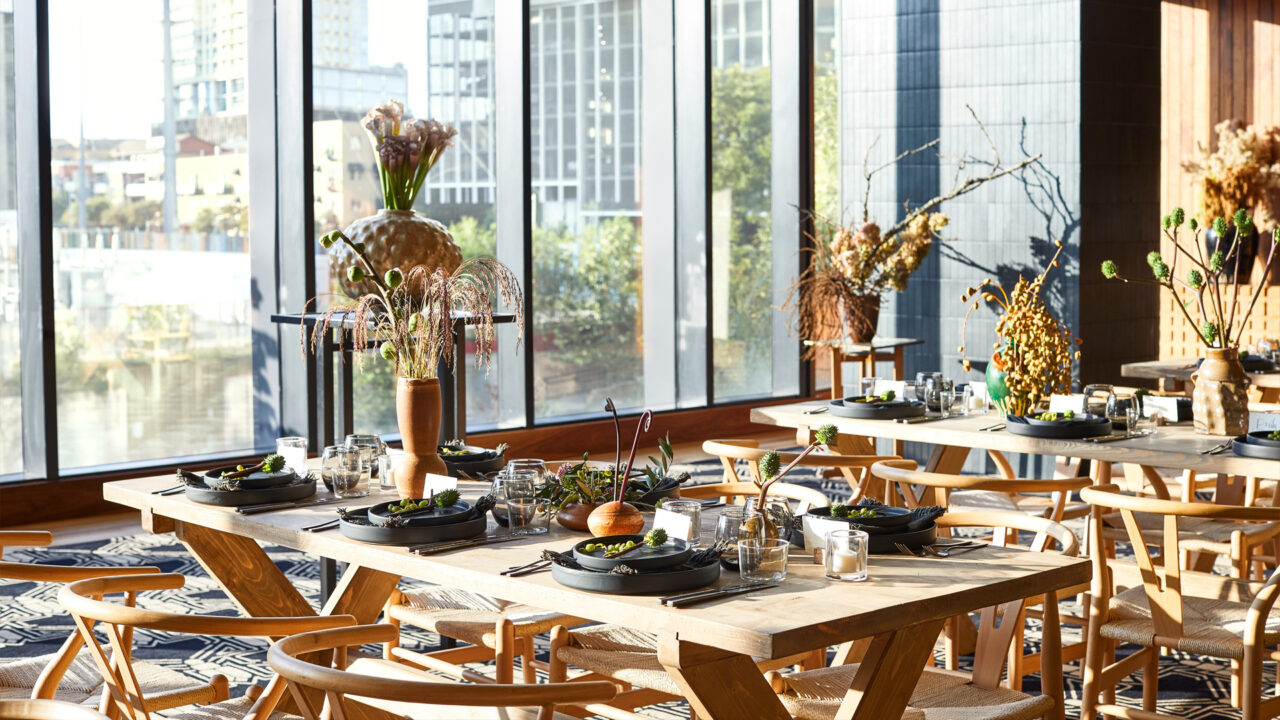 Sunlit dining area with wooden tables, modern decor, and large city-view windows.
