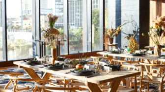 Sunlit dining area with wooden tables, modern decor, and large city-view windows.