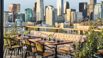 A rooftop dining area features a long table, sofas, and chairs, overlooking city skyscrapers.