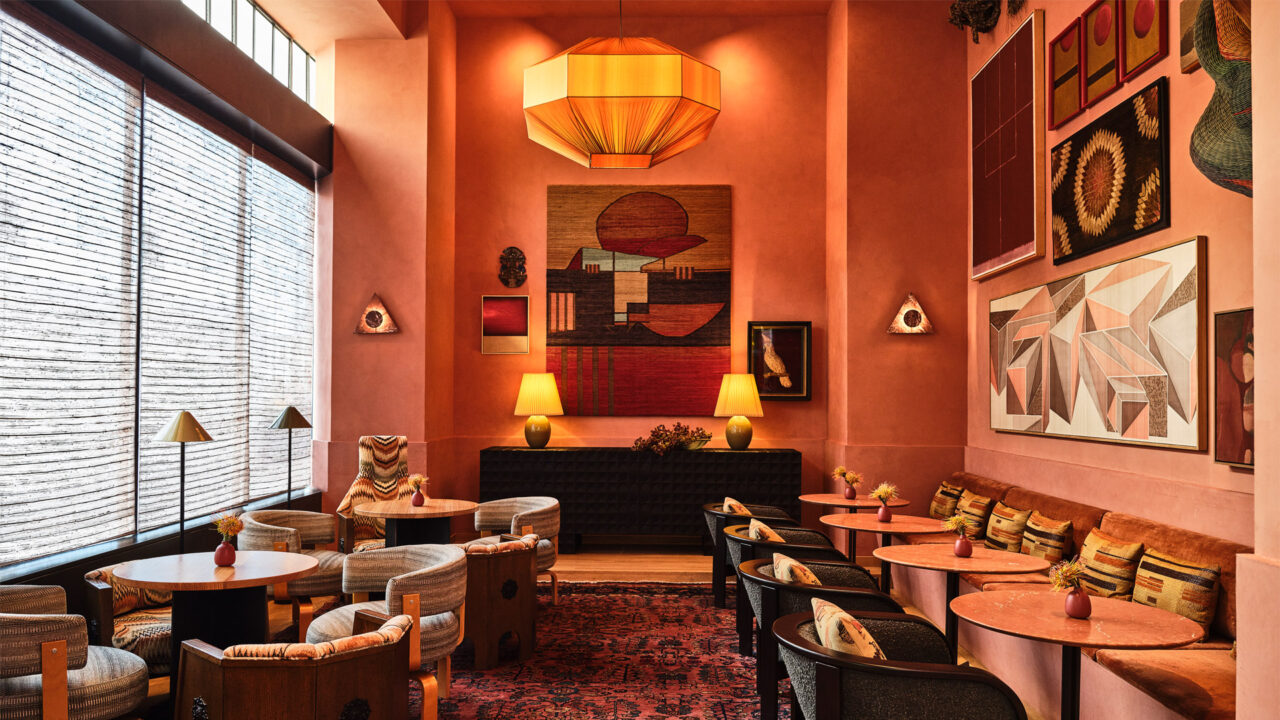 Warm-toned lounge with pink walls, eclectic artwork, round tables, and cozy seating under a large pendant light