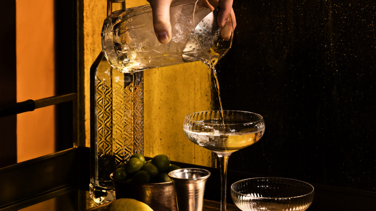 Bartender pouring a handcrafted cocktail beside olives and citrus in moody lounge lighting