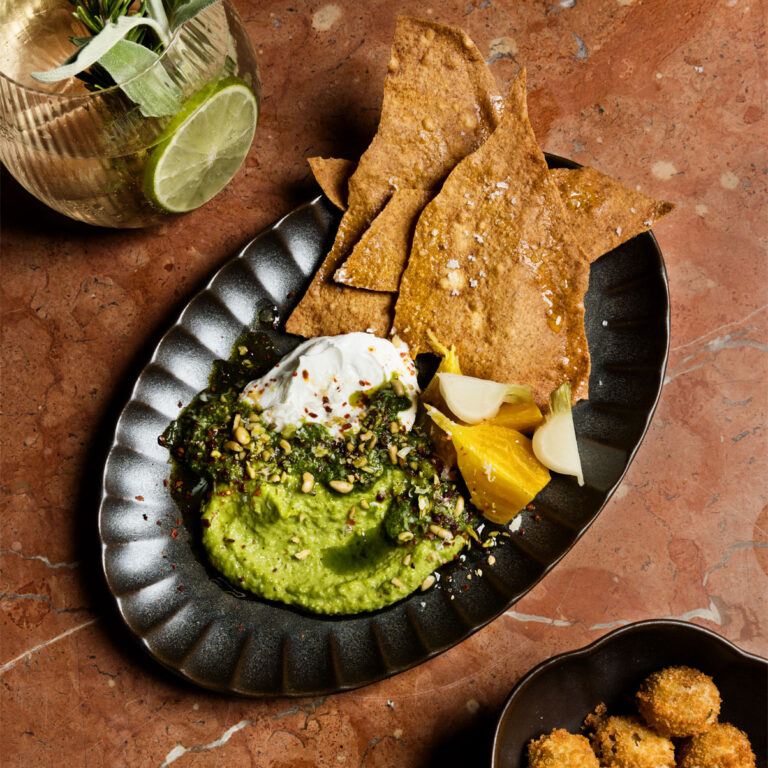 Plate of guacamole topped with herbs and seeds, served with crispy chips, pickled vegetables, and a lime cocktail on a stone table