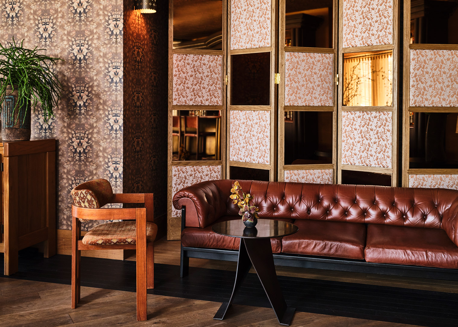 Cozy lounge with a tufted leather sofa, wooden chair, floral wallpaper, and decorative folding screens in warm ambient light