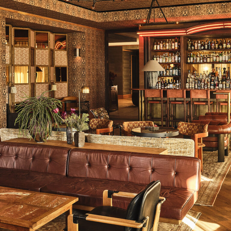 Cozy vintage lounge with leather sofa, wooden tables, patterned walls, and a backlit bar lined with bottles
