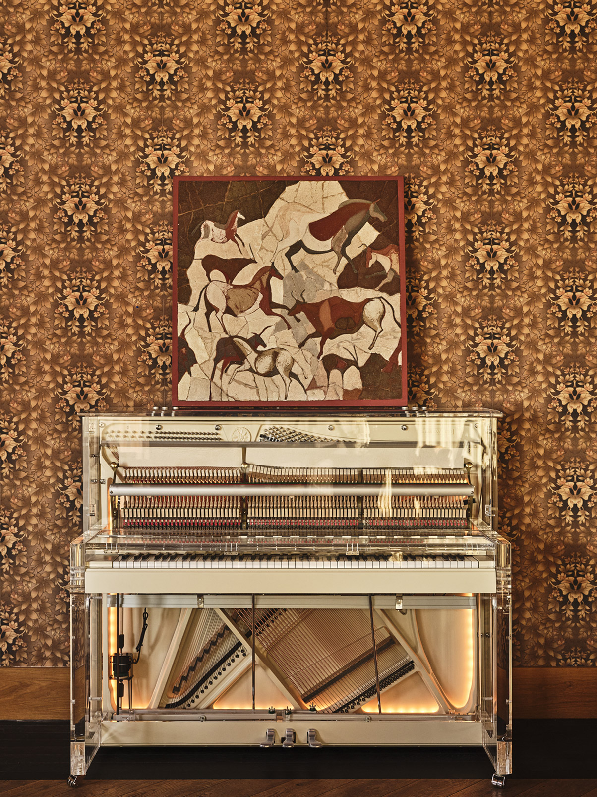 Transparent upright piano against patterned wallpaper with a framed artwork of abstract horses above