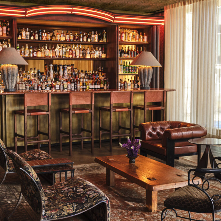 Elegant hotel bar with a backlit wall of liquor bottles, marble counter, leather stools, and warm ambient lighting