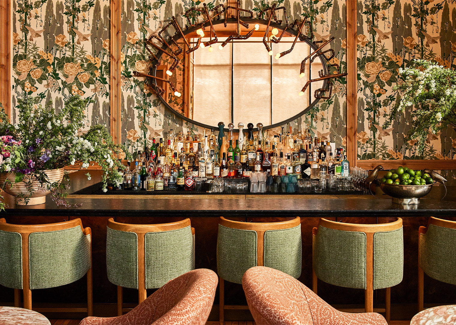 Elegant bar with floral wallpaper, circular mirror, and shelves of spirits and glassware