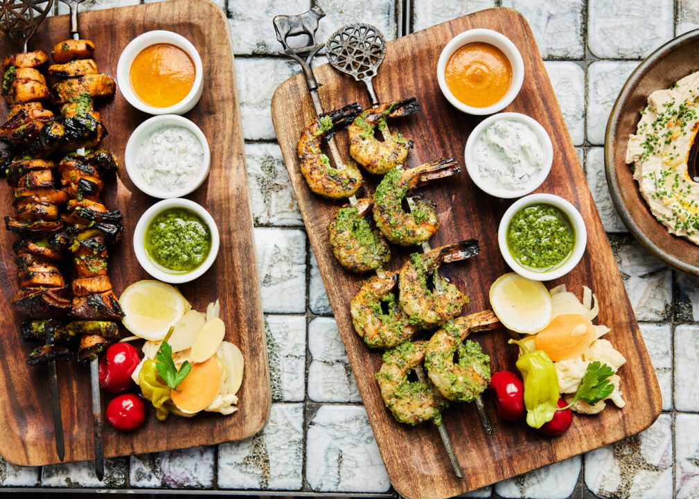 Grilled skewers and shrimp served on wooden boards with dipping sauces, lemon, and pickled vegetables