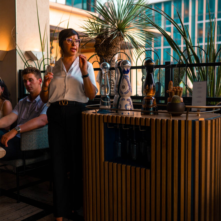 Woman holding a glass and presenting tequila bottles at an outdoor tasting setup