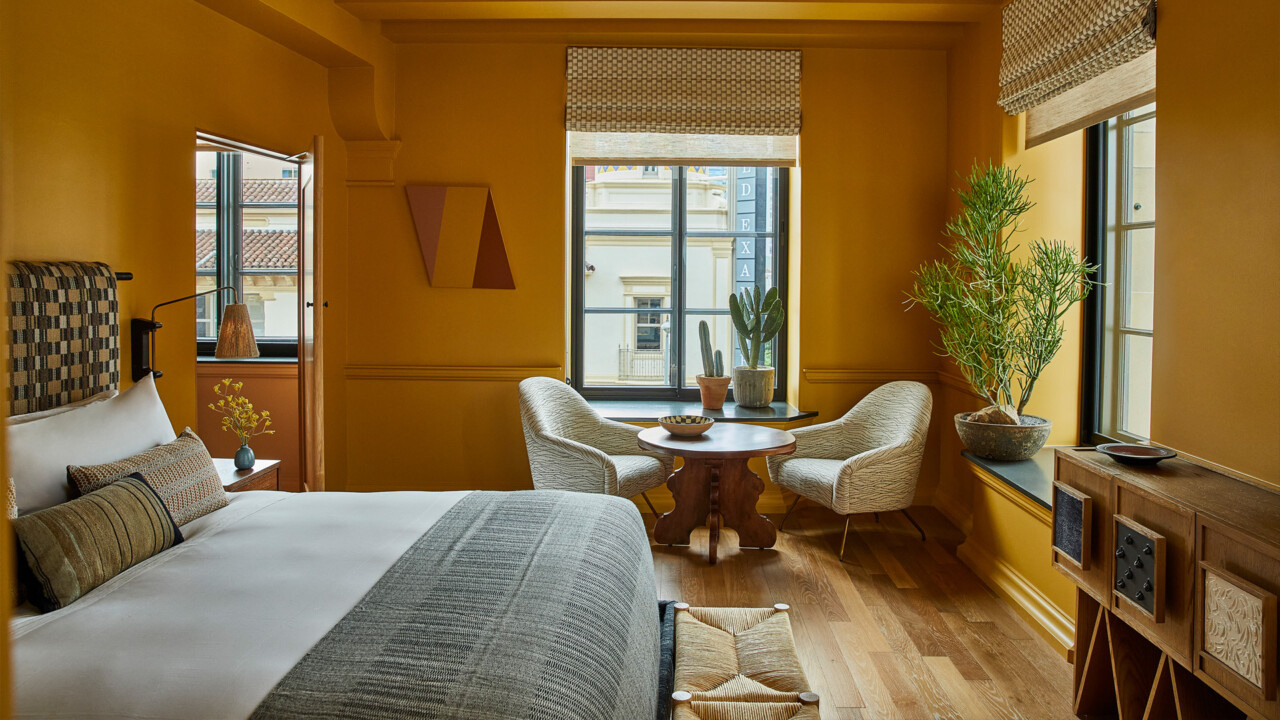 Warm-toned DTLA hotel room with a bed, cozy seating nook, and city views through large windows.