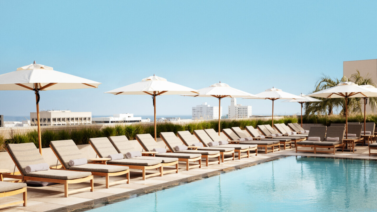 Rows of poolside loungers with white umbrellas reflected in turquoise water, overlooking a clear blue sky.