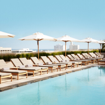 Rows of poolside loungers with white umbrellas reflected in turquoise water, overlooking a clear blue sky.