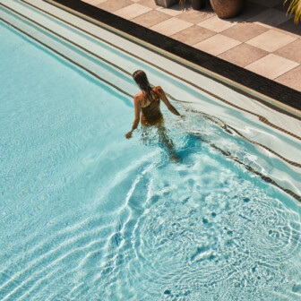 Woman in swimsuit wading through sunlit outdoor pool, clear blue water rippling around her legs.