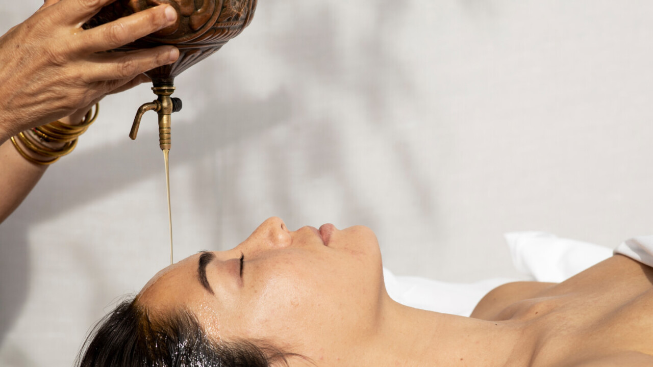 Oil is poured from brass vessel onto forehead of woman lying down, spa wellness treatment scene.