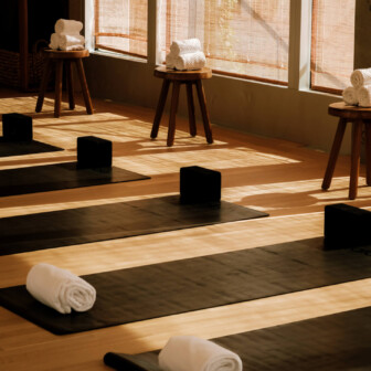Yoga mats with rolled towels, wooden blocks, and stools line sunlit studio floor near large windows.