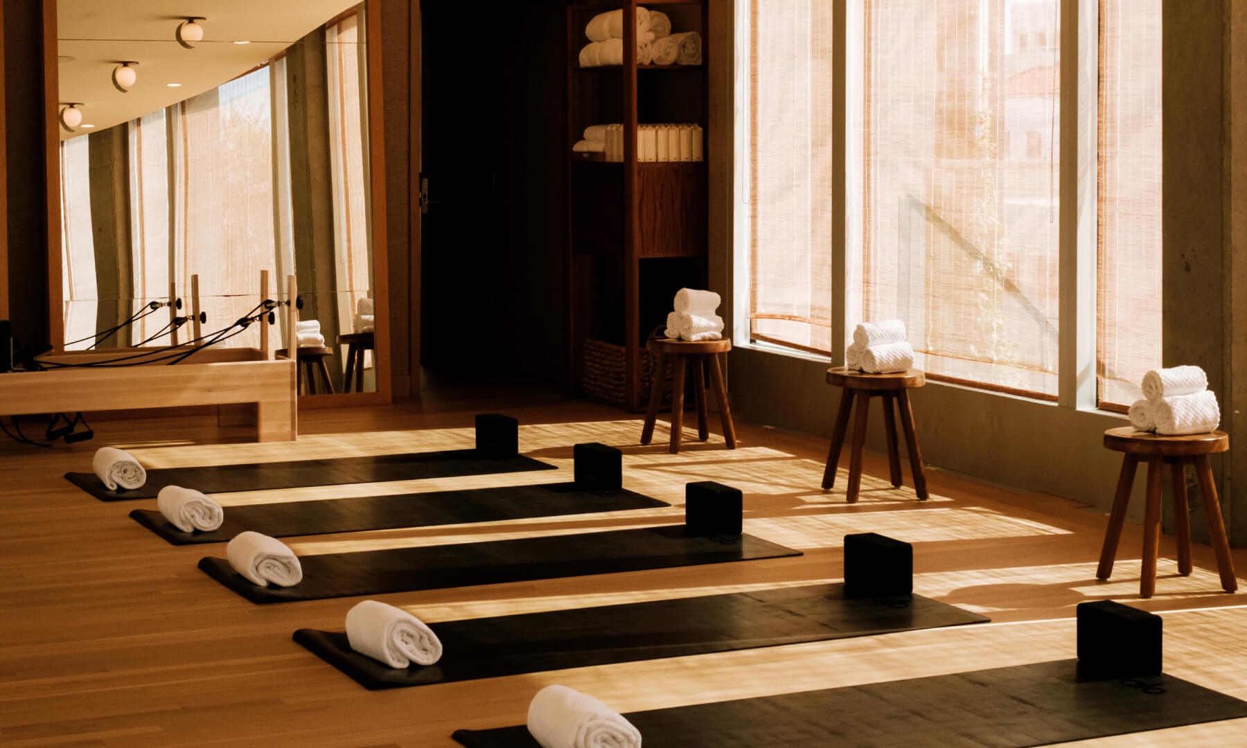 Yoga mats with rolled towels, wooden blocks, and stools line sunlit studio floor near large windows.