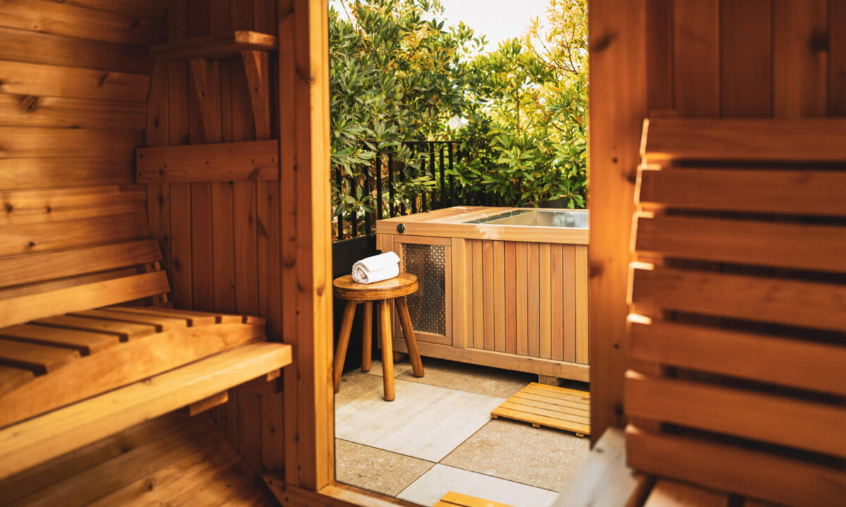 A wooden sauna interior opens to a patio with a hot tub, greenery, and a towel nearby.