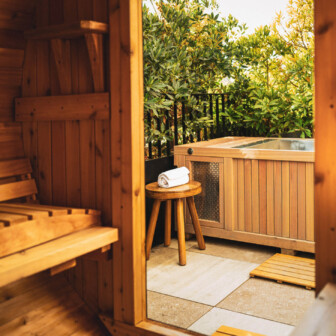 A wooden sauna interior opens to a patio with a hot tub, greenery, and a towel nearby.