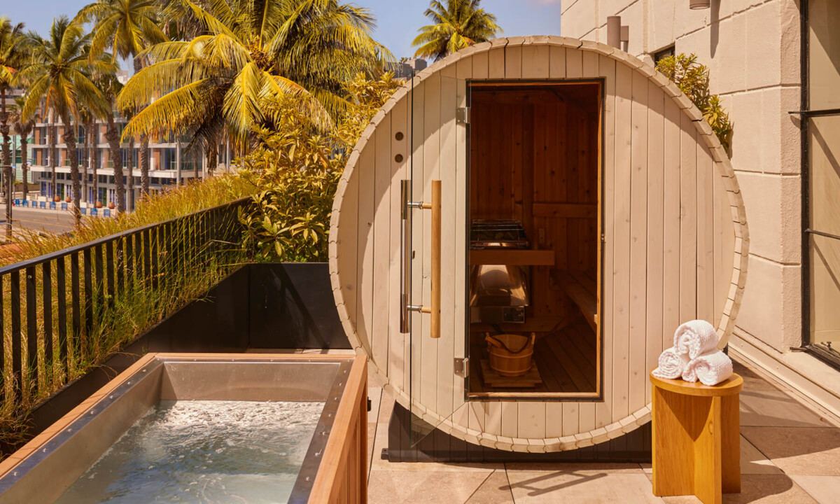 A round outdoor sauna with wooden door, nearby hot tub, palm trees, and towels stacked on stool.