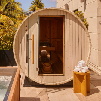 A round outdoor sauna with wooden door, nearby hot tub, palm trees, and towels stacked on stool.