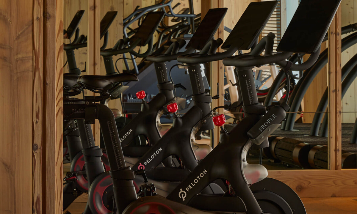 Stationary exercise bikes with digital screens are lined up in a wooden fitness room with warm lighting.