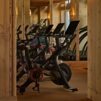 Stationary exercise bikes with digital screens are lined up in a wooden fitness room with warm lighting.