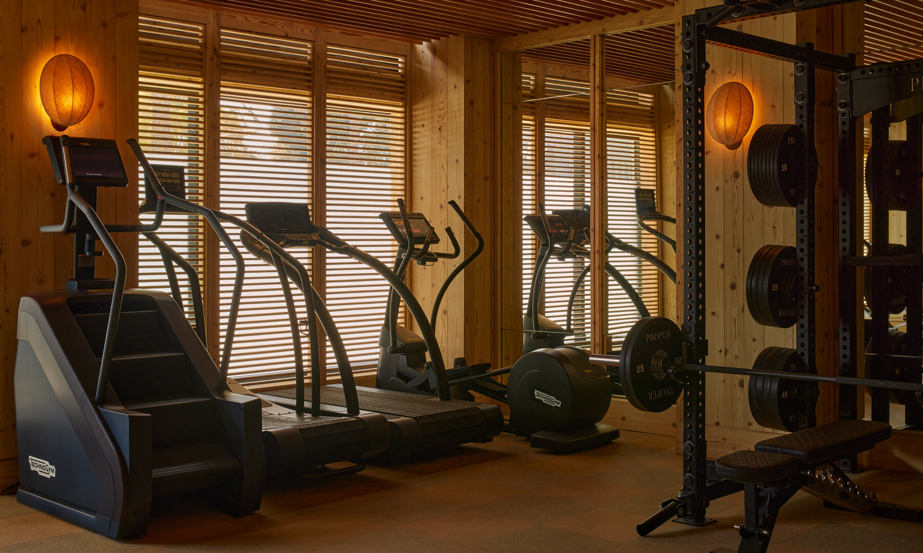 Modern gym with stair stepper, elliptical machines, and wood accents, sunlight filtered through window blinds.