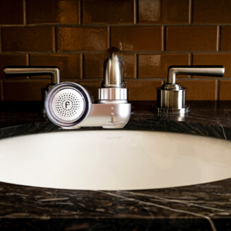 Modern bathroom sink with round faucet attachment and metallic handles, set against glossy brown tiled wall.