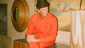 Person in bright orange jumpsuit and dark hat sitting on marble surface at Proper Hotel, smiling in front of decorative wall.