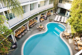 Curved outdoor pool surrounded by lounge chairs, dining tables, greenery, and multi-story hotel balconies above