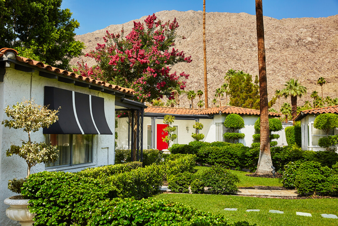 White casita homes with black awnings, lush manicured bushes, blooming pink tree, mountain backdrop, bright sunlight.