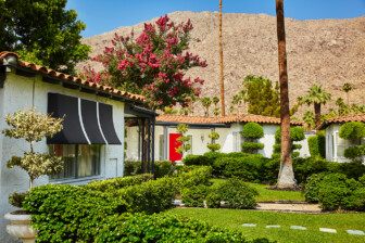 White casita homes with black awnings, lush manicured bushes, blooming pink tree, mountain backdrop, bright sunlight.