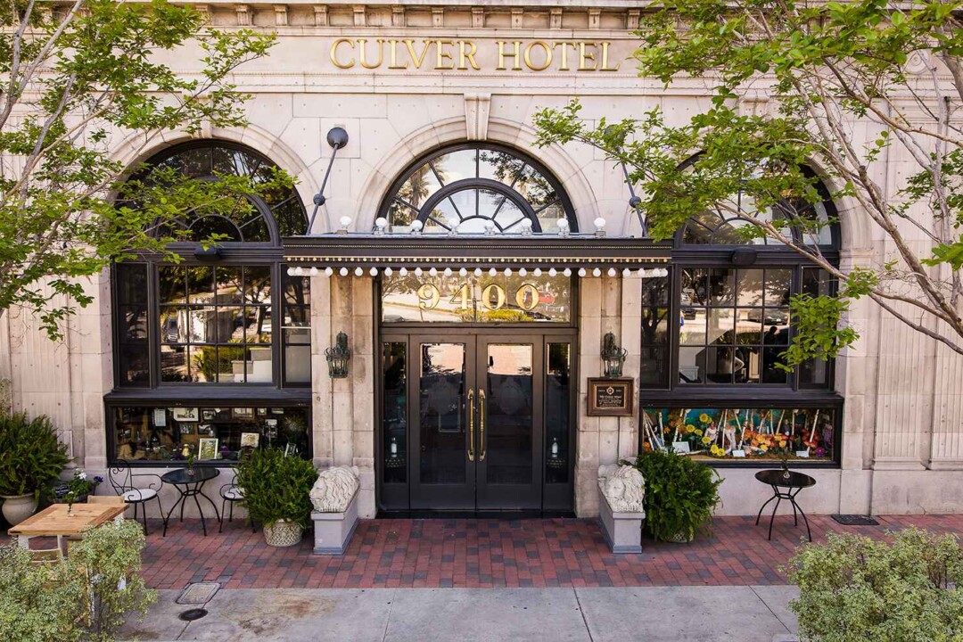 Historic Culver Hotel exterior with grand arched windows, black awning entrance, decorative columns, and leafy trees outside.