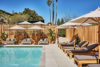 Outdoor swimming pool surrounded by wooden lounge chairs, white umbrellas, plants, and natural landscape under blue sky.