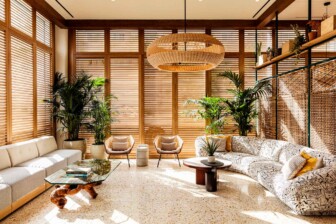 Sunlit lounge with large windows, natural wood blinds, patterned sofas, potted plants, and woven pendant light.